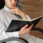 A man in traditional attire reads thoughtfully indoors, conveying contemplation.